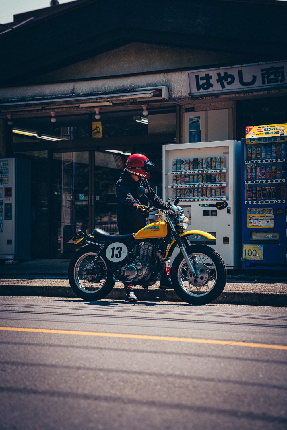 昔ながらの商店と自動販売機の前に停めた、黄色いSR400スクランブラー