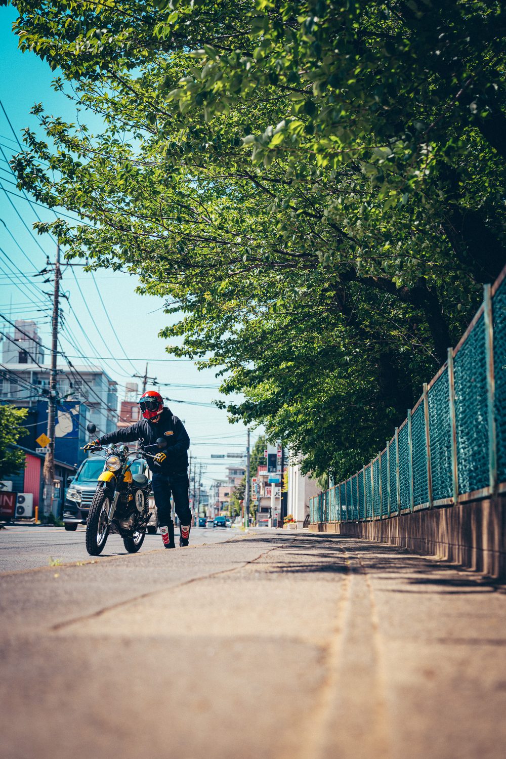 緑の木々に沿った街の歩道で、カスタムしたSR400を押すライダー