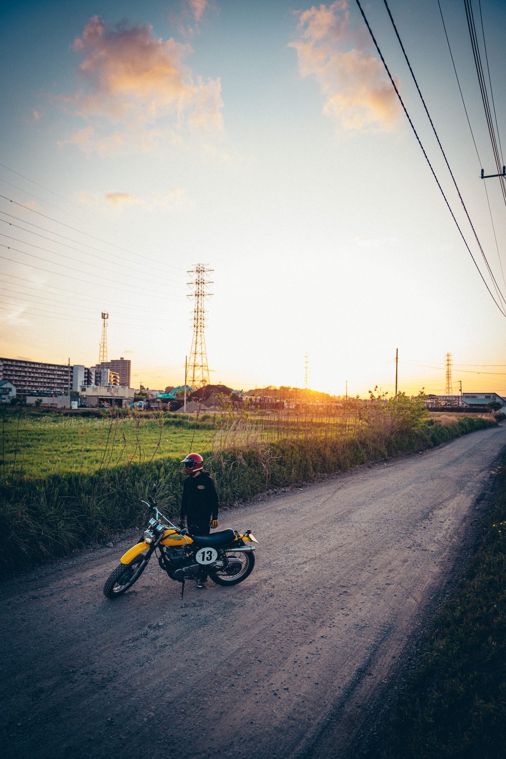 夕暮れの田畑に囲まれたダートに停めた、平成レトロなSR400スクランブラーとライダー